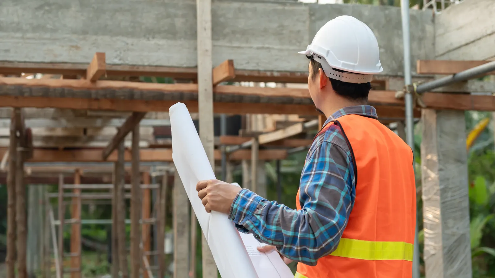 Builder in a hard hat and hi-vis vest holding rolled plans on a timber-framed site