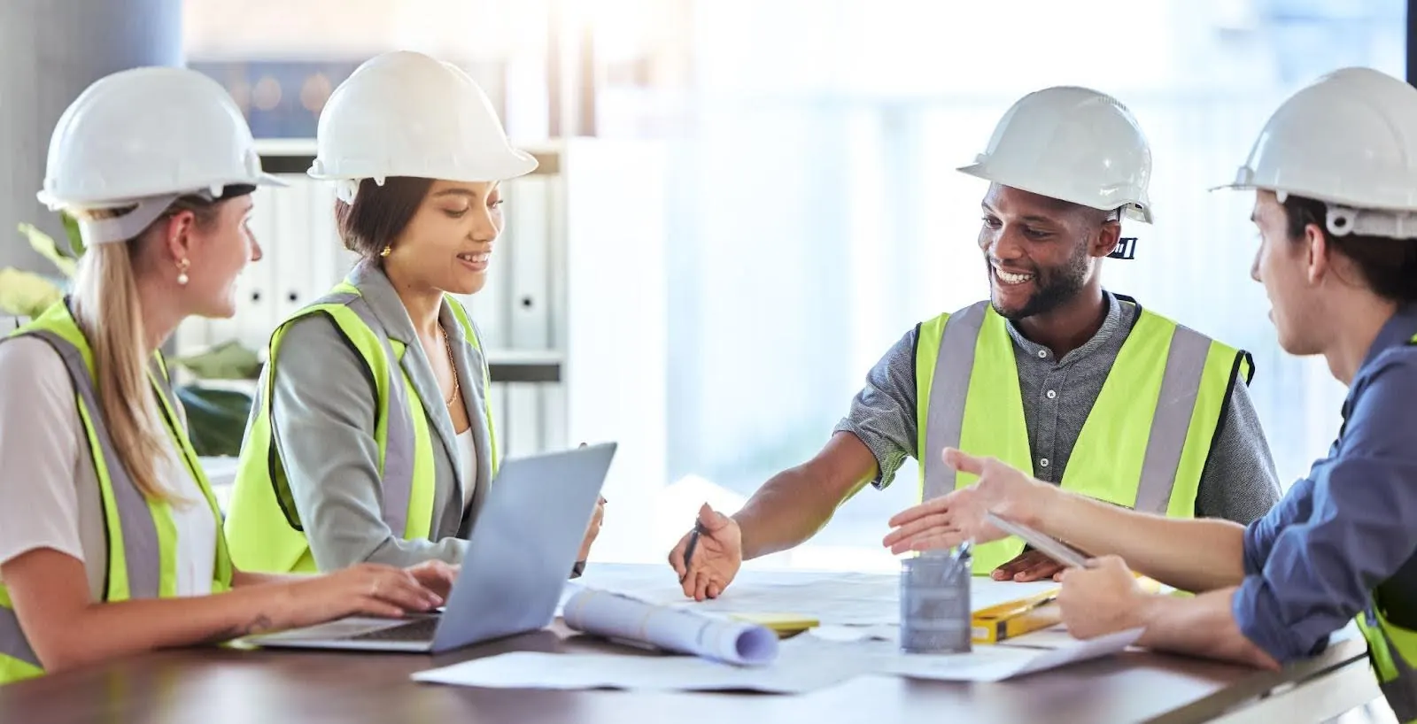 Construction team in hard hats and hi-vis vests meeting around a table with plans