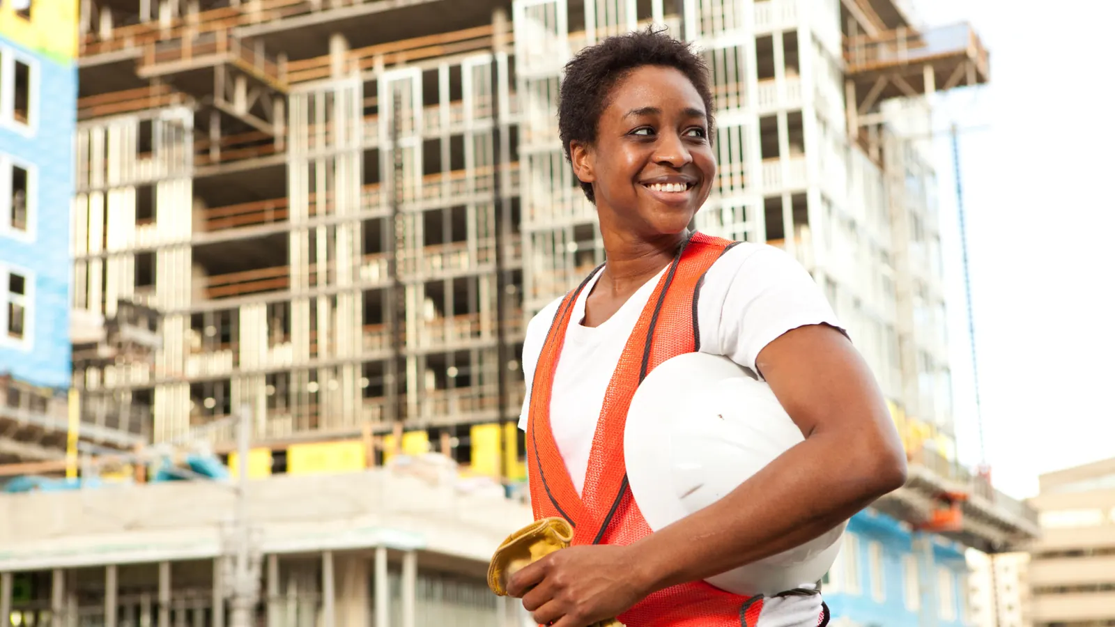 Smiling site worker in an orange vest holding a hard hat in front of a building under construction