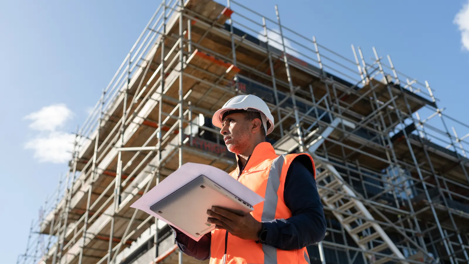 Builder holding a folder of plans in front of a scaffolded building